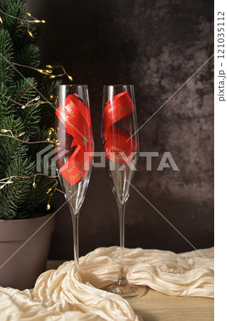 Christmas decor, two transparent glass with a red ribbon inside, tree with lights, dark background, beige cloth on wooden table Christmas decor, two transparent glass with a red ribbon inside, tree with lights, dark background, beige cloth on wooden table 121035112