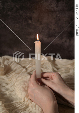 One white lit candle in woman hands on dark gray background, beige cloth 121035116