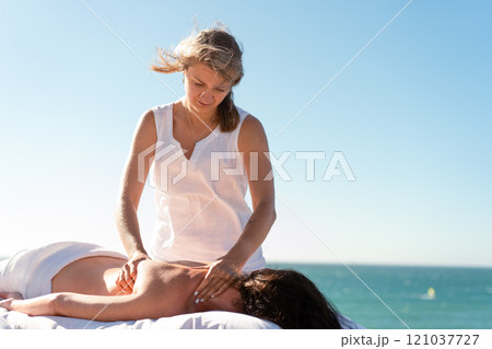 Therapist massaging woman's naked back against sea Therapist massaging woman's naked back against sea 121037727