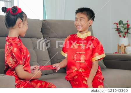 A happy sibling duo, dressed in red cheongsam qipao, exchange monetary gifts in hongbao envelopes at home. The living room scene captures the warmth and tradition of Chinese New Year A happy sibling duo, dressed in red cheongsam qipao, exchange monetary gifts in hongbao envelopes at home. The living room scene captures the warmth and tradition of Chinese New Year 121043360