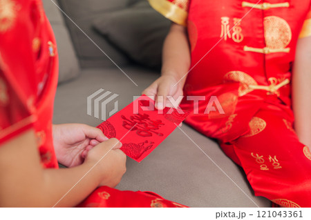 A happy sibling duo, dressed in red cheongsam qipao, exchange monetary gifts in hongbao envelopes at home. The living room scene captures the warmth and tradition of Chinese New Year A happy sibling duo, dressed in red cheongsam qipao, exchange monetary gifts in hongbao envelopes at home. The living room scene captures the warmth and tradition of Chinese New Year 121043361