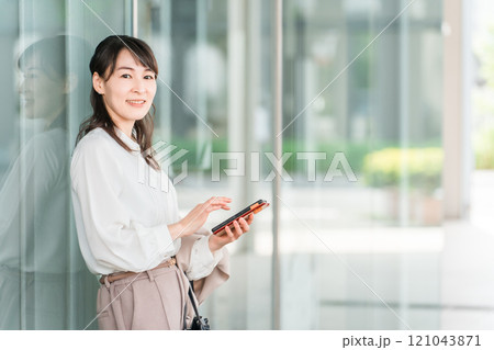 オフィスビル・商業施設の前でスマホを見ながら待ち合わせするビジネスウーマン・ワーキングマザー 121043871