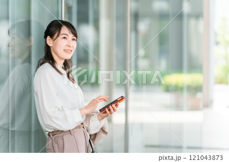 オフィスビル・商業施設の前でスマホを見ながら待ち合わせするビジネスウーマン・ワーキングマザー オフィスビル・商業施設の前でスマホを見ながら待ち合わせするビジネスウーマン・ワーキングマザー 121043873
