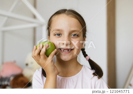 Little Caucasian girl with missing teeth from falling out milk teeth smiling and holding apple indoors, conveying joy and childhood innocence. Little Caucasian girl with missing teeth from falling out milk teeth smiling and holding apple indoors, conveying joy and childhood innocence. 121045771