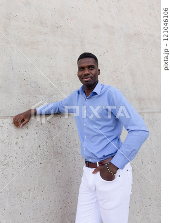 Portrait Of Handsome Smiling Tall African American Man wearing White Jeans and Blue Shirt, Brown Belt Outdoor, Concrete Wall on Background. Copy Space Fashionable Male, Ivorian. Vertical Plane Portrait Of Handsome Smiling Tall African American Man wearing White Jeans and Blue Shirt, Brown Belt Outdoor, Concrete Wall on Background. Copy Space Fashionable Male, Ivorian. Vertical Plane 121046106