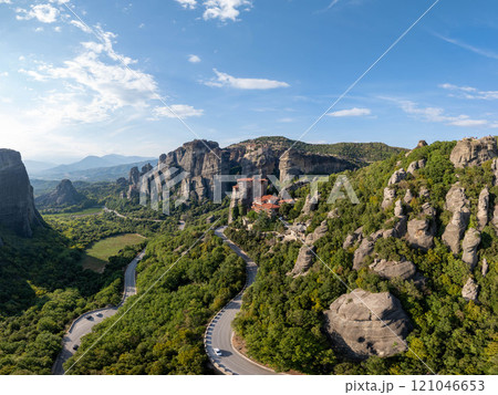 Cars navigating through winding roads amidst the stunning scenery of Meteora, Greece, with towering rock formations and a historic monastery creating a picturesque backdrop 121046653