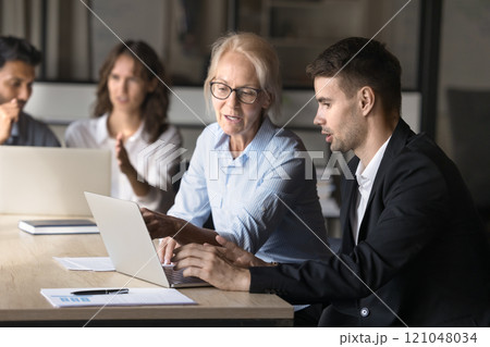 Elderly woman explains tasks on laptop to younger colleague 121048034