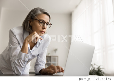 Thoughtful grey haired freelancer woman in glasses looking at laptop Thoughtful grey haired freelancer woman in glasses looking at laptop 121048175