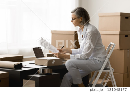 Serious storage employee woman reading parcel card at workplace Serious storage employee woman reading parcel card at workplace 121048177