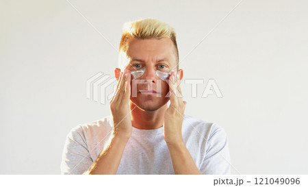 Eye patches. Anti-aging treatment. Confident blond man applying moisturizing collagen hydrogel pads on clean fresh face skin isolated on white empty space. Eye patches. Anti-aging treatment. Confident blond man applying moisturizing collagen hydrogel pads on clean fresh face skin isolated on white empty space. 121049096
