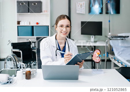 Young female doctor in white medical uniform using laptop and tablet talking video conference call at desk,Doctor sitting at desk and writing a prescription for her patient 121052865