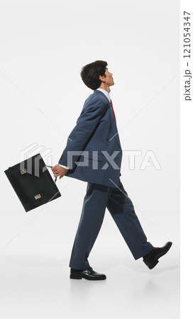 Confident businessman in blue suit and red tie walking dynamically while holding black briefcase against white background. 121054347