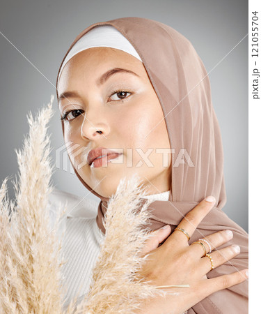 Studio portrait of one beautiful young muslim woman wearing brown headscarf posing with pampas wheat plant against grey background. Modest arab wearing natural makeup and covered in traditional hijab 121055704