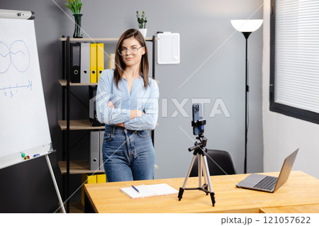Young woman mentor recording presentation, webinar on smartphone standing on tripod, teacher coach explaining, holding online lesson, using phone webcam. Young woman mentor recording presentation, webinar on smartphone standing on tripod, teacher coach explaining, holding online lesson, using phone webcam. 121057622