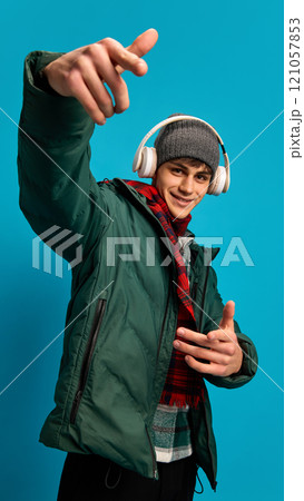 Young smiling man wearing winter puff jacket, red checkered scarf and gray hat, listening to music in headphones against blue studio background 121057853