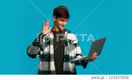 Portrait of handsome young smiling man holding laptop and waving towards screen online communication via video call, against blue studio background 121057858