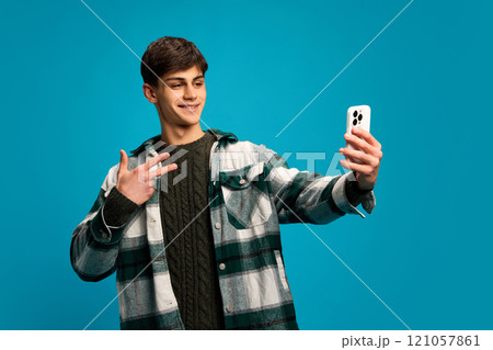 Handsome young guy in casual clothes, checkered shirt taking selfie with smartphone against blue studio background. Social media, communication 121057861
