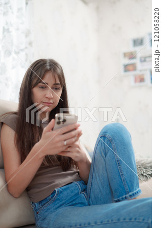 Young happy woman sitting comfortably on sofa, smiling while using her phone indoors during a relaxing afternoon vertical Young happy woman sitting comfortably on sofa, smiling while using her phone indoors during a relaxing afternoon vertical 121058220