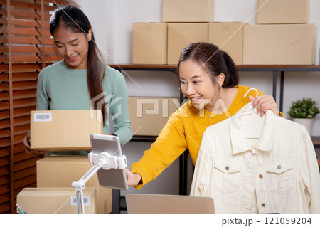 Two young asian woman live online clothing store while showing clothing with tablet and laptop. 121059204