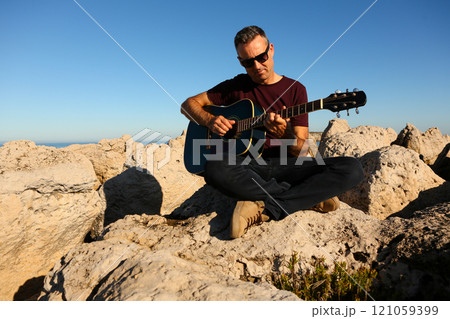 A man in a dark t-shirt playing acoustic guitar on a rocky area by the sea 121059399