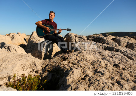 A man in a dark t-shirt playing acoustic guitar on a rocky area by the sea 121059400