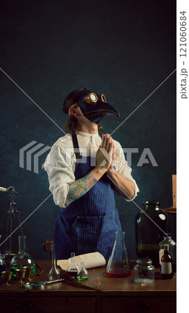 Mysterious m in plague doctor mask stands in vintage lab, hands clasped in prayer-like gesture, surrounded by flasks and glassware on wooden table. 121060684
