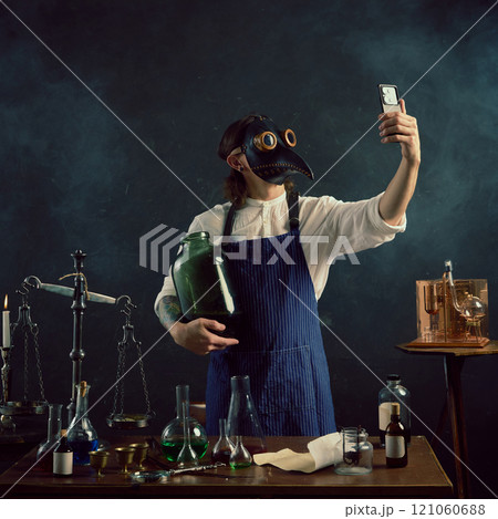 Holding large glass jar, the man in plague doctor mask takes modern selfie, hinting at his unconventional brewing or distilling work. 121060688