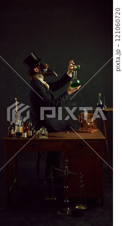 Plague doctor mixing his elements, experimenting and researching in vintage laboratory against dark mystery background. 121060720