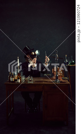 Plague doctor in traditional attire examines mysterious vial, surrounded by array of antique medical tools and glass containers in vintage laboratory Plague doctor in traditional attire examines mysterious vial, surrounded by array of antique medical tools and glass containers in vintage laboratory 121060724