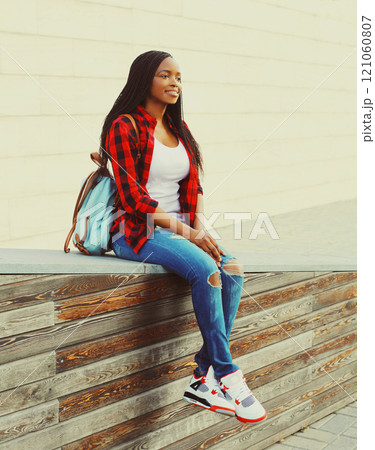 Fashion smiling african woman wearing a red checkered shirt and backpack resting in the city park 121060807