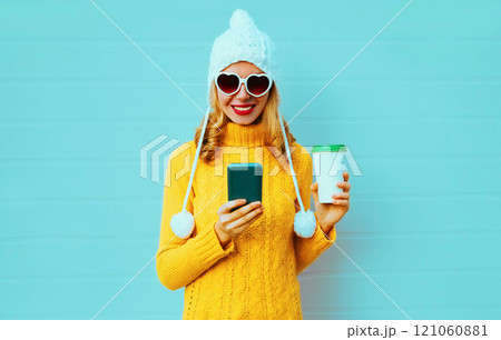 Winter portrait smiling young woman with phone and coffee cup in yellow sweater, white hat on blue Winter portrait smiling young woman with phone and coffee cup in yellow sweater, white hat on blue 121060881