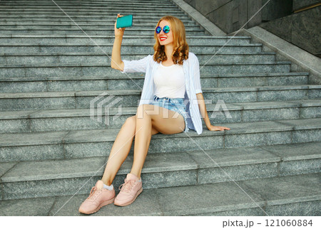 Happy smiling young woman taking selfie picture by smartphone sitting on the stairs on city street 121060884