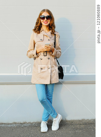 Fashion lovely young smiling woman using smartphone wearing a coat handbag clutch in city 121060889
