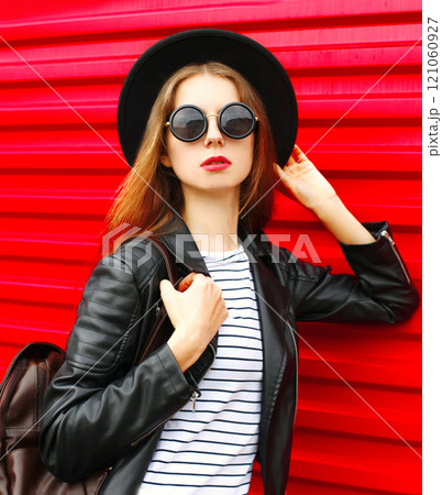 Fashion portrait young woman in black rock style hat, jacket stands with backpack on red background 121060927