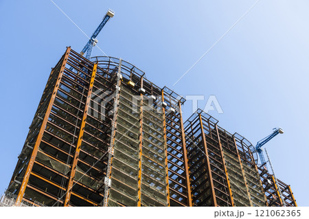 Low-angle view of cranes and steel structures of building construction with a blue sky background. Low-angle view of cranes and steel structures of building construction with a blue sky background. 121062365