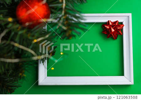A white wooden frame lies in the center of the image, with a red bow in the right corner and on the left of the image you can see the branches of a Christmas tree with a red shiny ball and a yellow 121063508