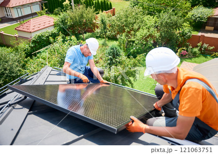 Workers building photovoltaic solar panel system on rooftop of house. Men technicians in helmets and gloves installing solar module with help of hex key outdoors. Renewable energy generation concept. Workers building photovoltaic solar panel system on rooftop of house. Men technicians in helmets and gloves installing solar module with help of hex key outdoors. Renewable energy generation concept. 121063751