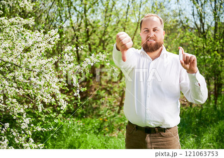 Man allergic using medical nasal spray, suffering from seasonal allergy at spring in blossoming garden. Handsome smiling man showing thumbs down near blooming tree outdoors. Spring allergy concept. 121063753