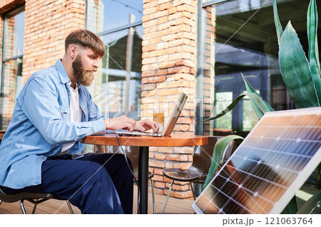 Man works on laptop at outdoor round table, with glass of iced coffee. Solar panel nearby, emphasizing sustainable, modern eco-friendly workspace that combines technology and sustainability. 121063764