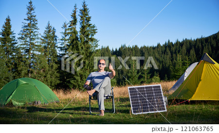Man using photovoltaic solar panel for charging portable power station outdoor in eco-friendly camping. Male tourist giving thumbs up near tents. On background tall pine trees and clear blue sky. Man using photovoltaic solar panel for charging portable power station outdoor in eco-friendly camping. Male tourist giving thumbs up near tents. On background tall pine trees and clear blue sky. 121063765