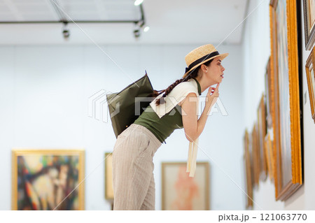 Mid shot of a young Caucasian woman in a straw hat and elegant clothes carefully contemplating modern art in the gallery. Low angle side view Mid shot of a young Caucasian woman in a straw hat and elegant clothes carefully contemplating modern art in the gallery. Low angle side view 121063770