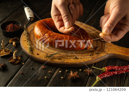 A skilled artisan carefully ties fresh sausage on a handcrafted wooden board. Surrounding ingredients include vibrant spices and herbs, creating an inviting culinary atmosphere A skilled artisan carefully ties fresh sausage on a handcrafted wooden board. Surrounding ingredients include vibrant spices and herbs, creating an inviting culinary atmosphere 121064209