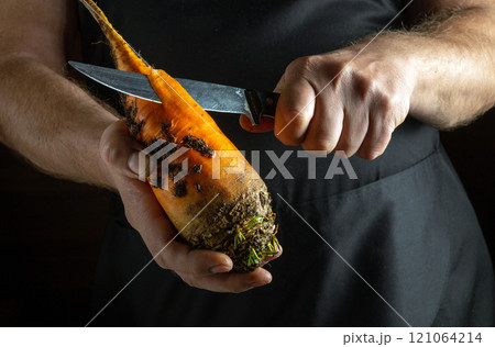 Focused hands grip a bright orange carrot, skillfully peeling away the skin with a sharp knife. The rustic kitchen ambiance adds warmth to the creative process of preparing fresh ingredients 121064214