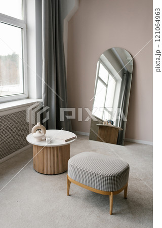 Small room with a white table and a wooden ottoman. Mirror is on the wall Small room with a white table and a wooden ottoman. Mirror is on the wall 121064963