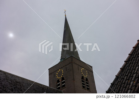 A tall church steeple with a clock and a weather vane stands against a cloudy gray sky, partially surrounded by rooflines, creating a serene and timeless architectural scene. A tall church steeple with a clock and a weather vane stands against a cloudy gray sky, partially surrounded by rooflines, creating a serene and timeless architectural scene. 121066802