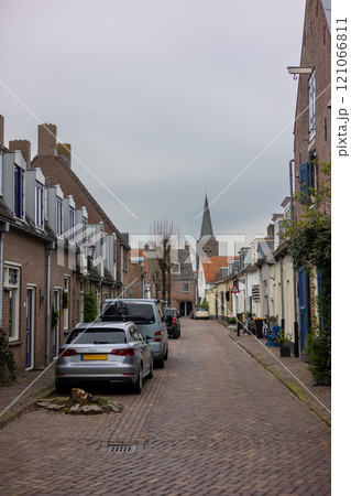 A quiet cobblestone street lined with historic brick buildings, adorned with shutters, parked cars, and traditional streetlights under an overcast sky. 121066811