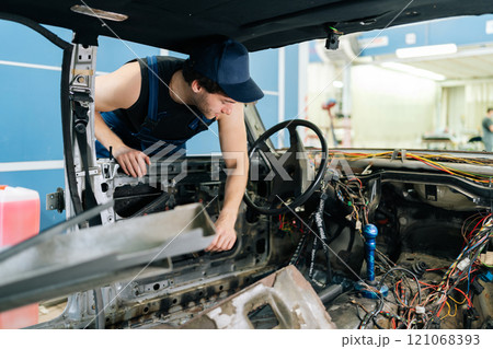 View from interior of old vehicle to mechanic in blue overalls inspecting car wiring after removing dashboard, showing expertise in troubleshooting automotive problems. Concept of car repair 121068393