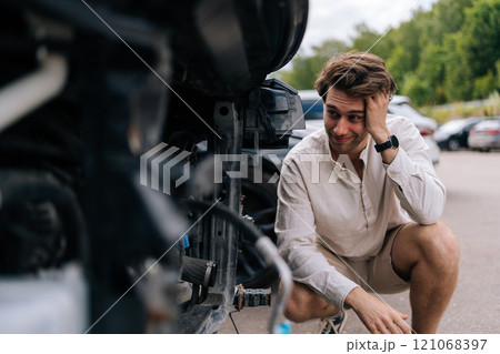 Unhappy young driver man assesses damage to car following accident on street, concerned about expenses for repairs and insurance. Pensive car owner crouching next to damaged car in parking lot. Unhappy young driver man assesses damage to car following accident on street, concerned about expenses for repairs and insurance. Pensive car owner crouching next to damaged car in parking lot. 121068397