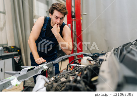 Portrait of pensive mechanic having stressful day at car repair shop, looking at car engine. Confused repairman unsure of how to fix complex engine problem in garage. Concept of car service, repair. 121068398