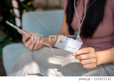 A close-up of an Asian woman sits on a sofa indoors, showing her credit card to the camera. 121072112